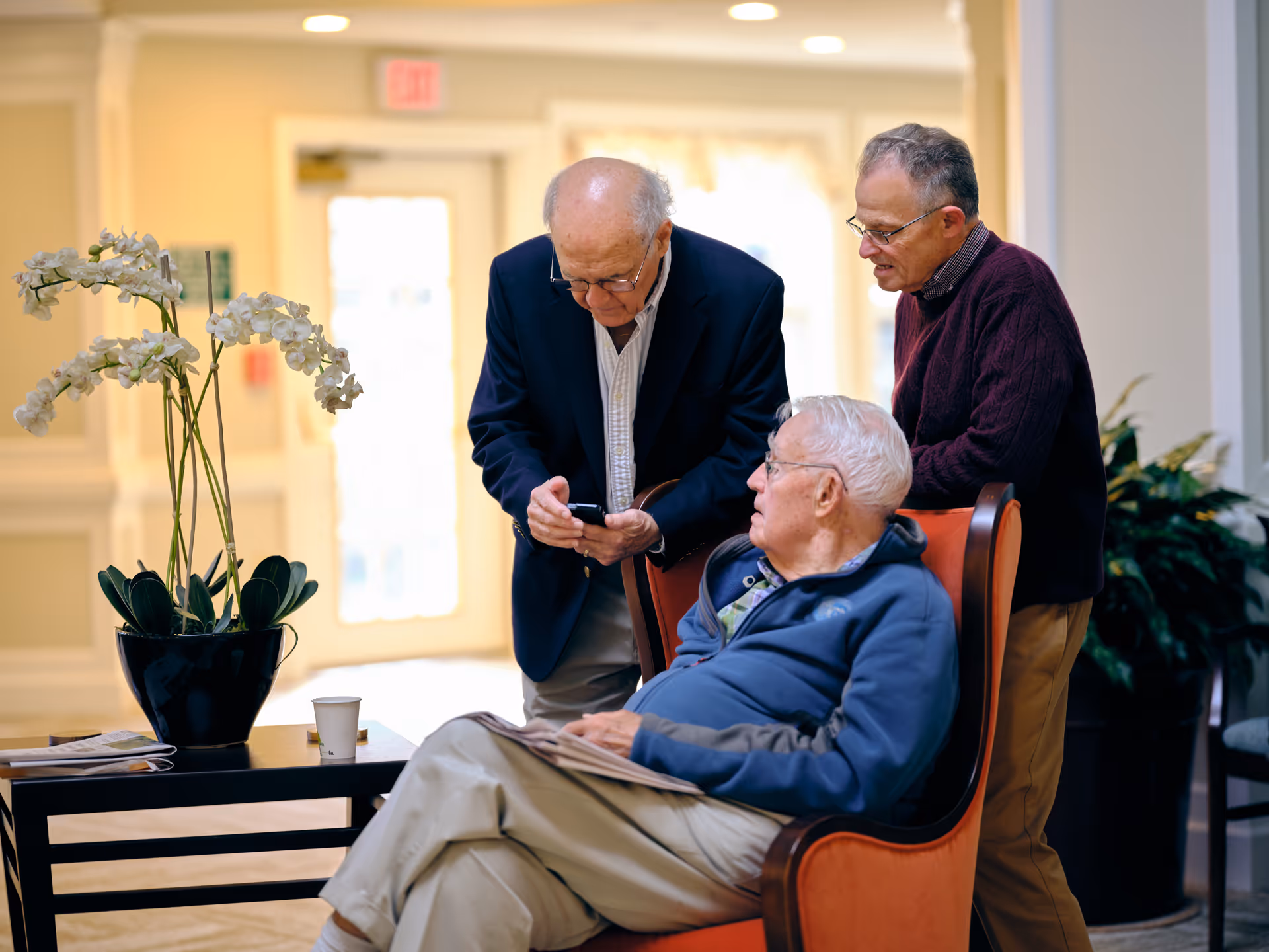 Three elderly men in a well-lit indoor common area. One man is seated in an orange armchair holding a newspaper, while the other two men stand nearby, one showing something on his phone to the others. There is a table with a potted orchid plant and a paper cup next to them.