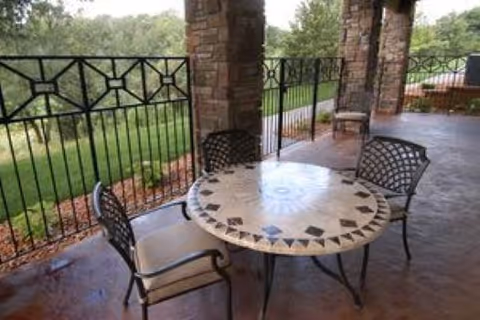 Covered outdoor patio with a round mosaic table and four metal chairs beside stone pillars and a decorative railing overlooking greenery.