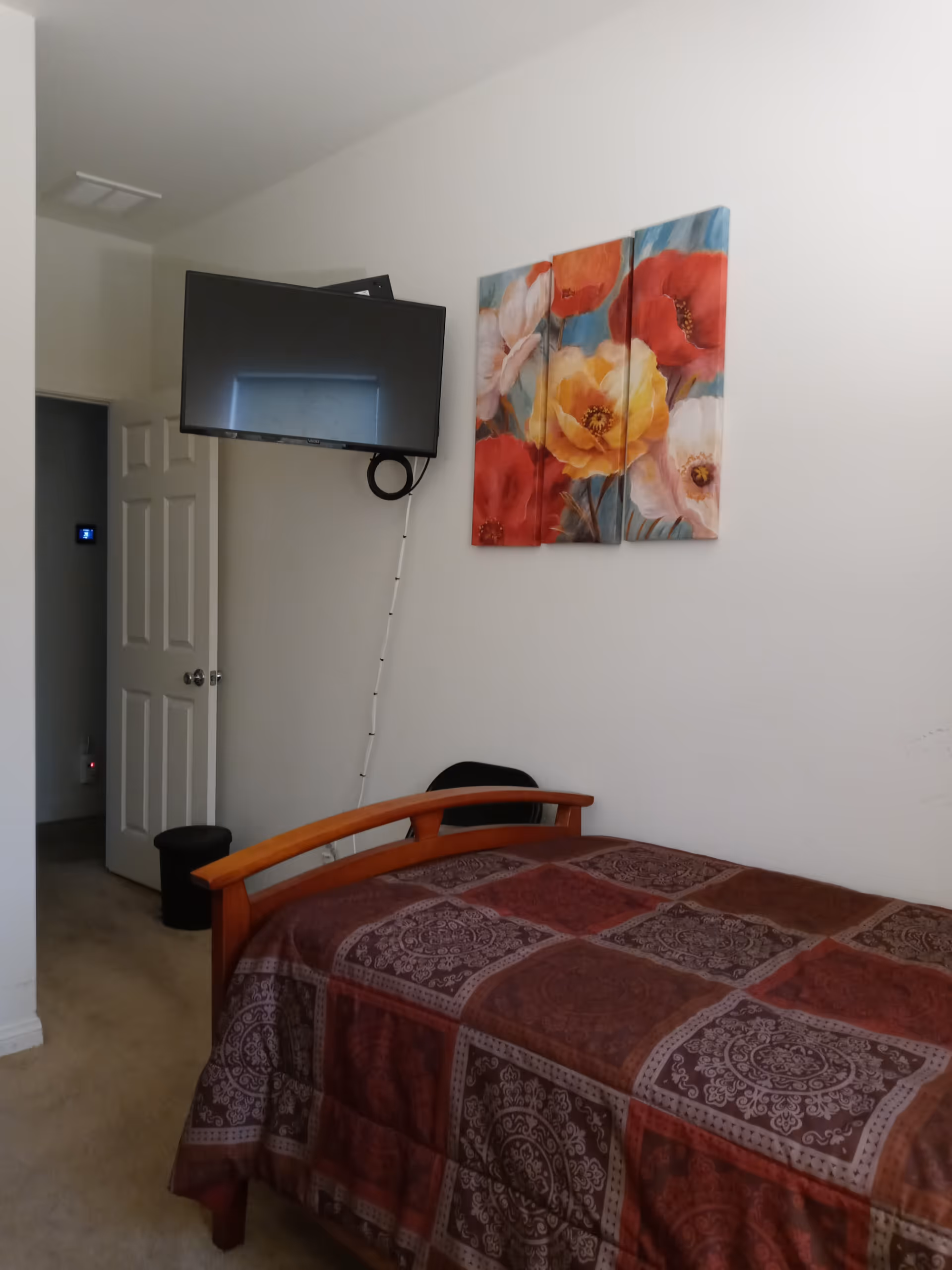 A bedroom with a single wooden bed covered with a patterned brown and red bedspread. Above the bed, there is a three-panel floral painting with red, yellow, and white flowers. A flat-screen TV is mounted on the wall to the left of the painting. An open door leads to another room, and a black trash bin is visible near the door.