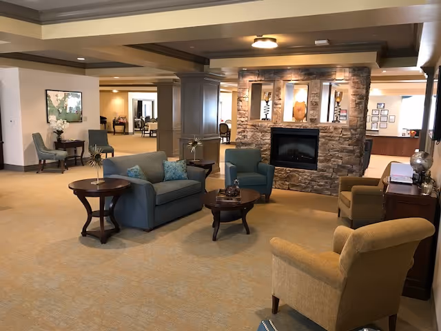 A cozy living room area in a senior living facility with a stone fireplace as the focal point. The room features comfortable seating including a blue loveseat, a blue armchair, and two beige armchairs arranged around wooden coffee and side tables. The space is well-lit with ceiling lights and has neutral-colored walls and carpet. In the background, there are additional seating areas and a reception desk.
