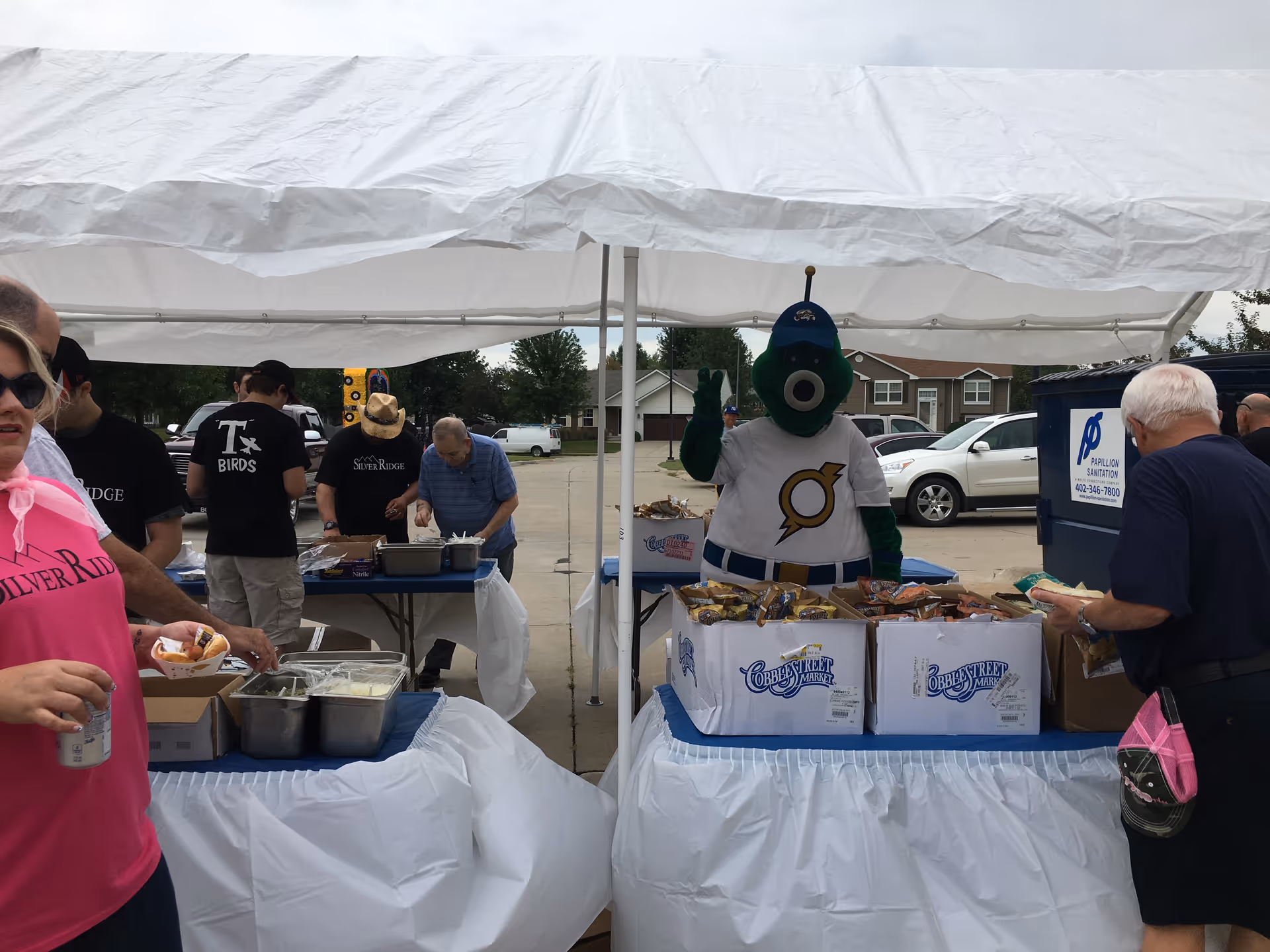 Outdoor event at Hillcrest Silver Ridge with people gathered under a white canopy tent. A person in a green mascot costume wearing a white baseball jersey and blue cap is standing behind a table with boxes of snacks. Other people are serving or getting food from tables covered with white tablecloths. Cars and houses are visible in the background.