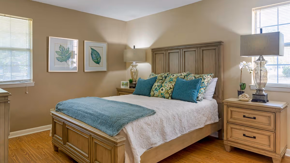 A furnished bedroom with a wooden bed dressed in white linens and teal pillows and throw, flanked by nightstands with lamps and framed leaf artwork on the wall.