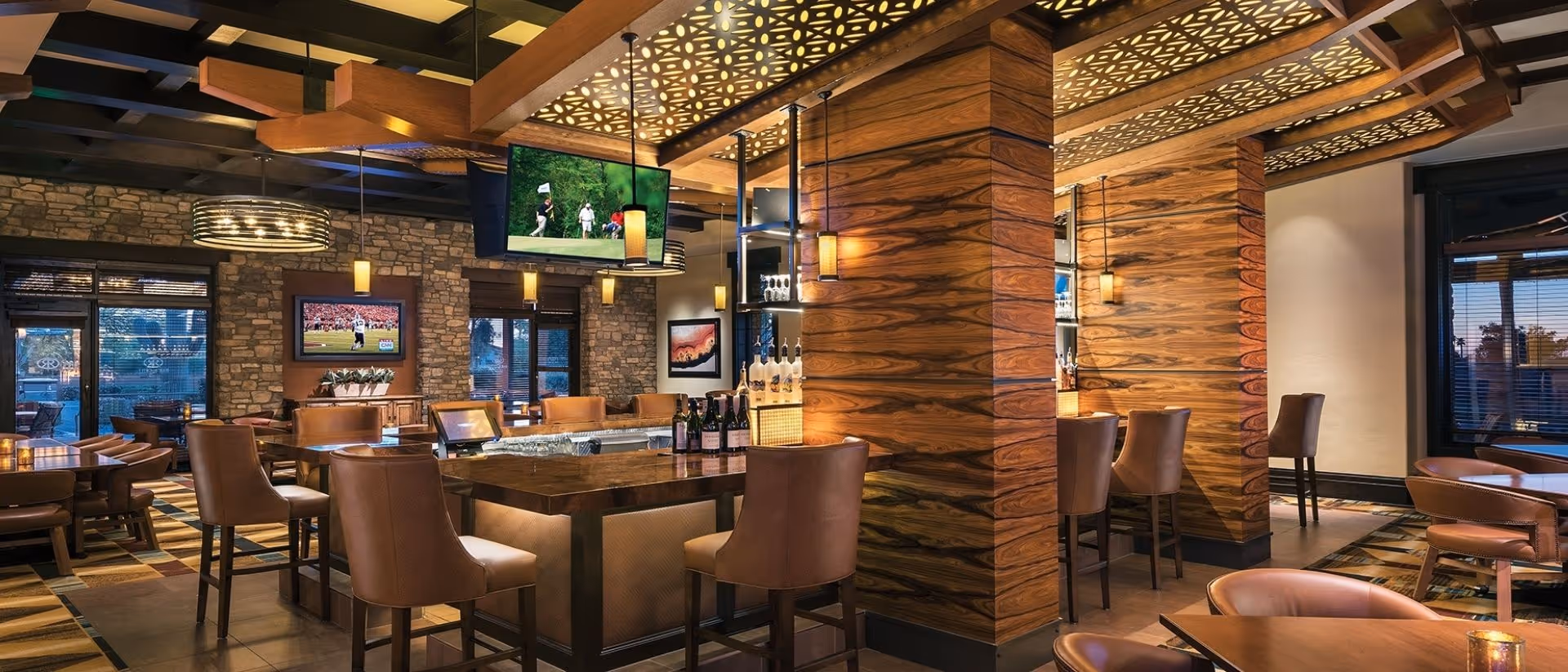 A stylish wood-accented bar and dining area with leather chairs, a central island bar, hanging televisions, and warm pendant lighting.