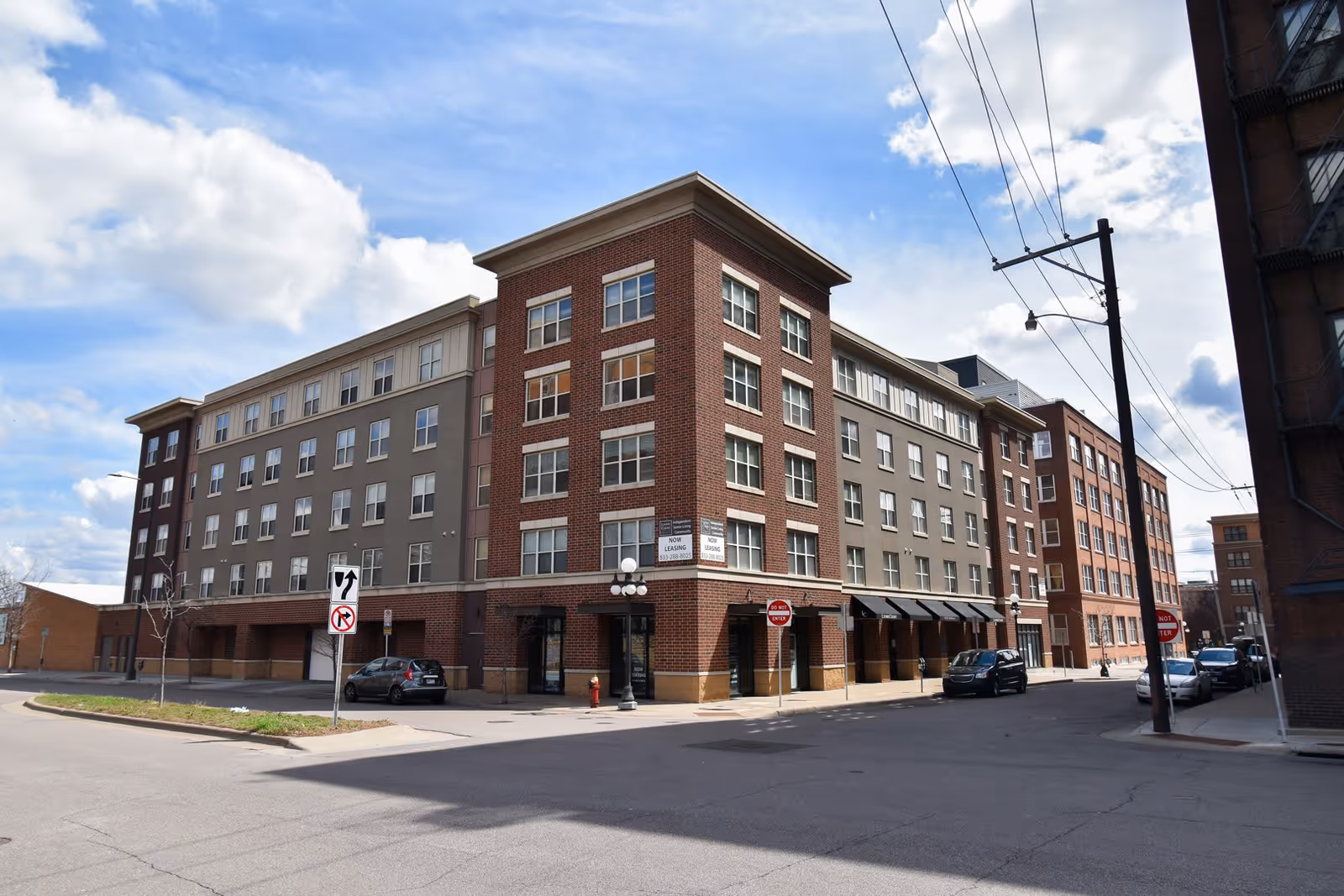 Exterior view of a multi-story senior apartment building named Lyons Court Senior Apartments, featuring a combination of brick and gray facade with multiple windows, located at a street corner under a partly cloudy sky.