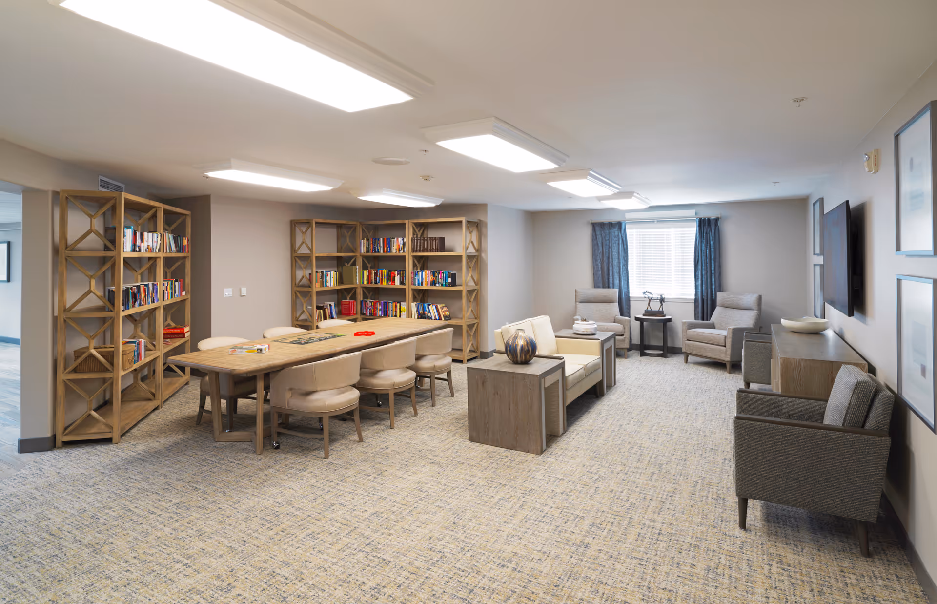 A spacious and well-lit common area in a senior living facility featuring a long wooden table with beige cushioned chairs, bookshelves filled with books, a small seating area with armchairs and a loveseat, a window with blue curtains, and a wall-mounted television.