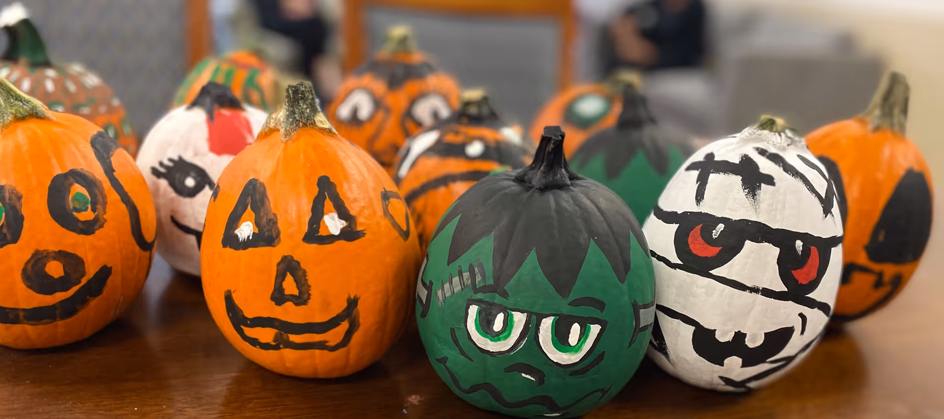 A collection of small pumpkins painted with various Halloween-themed faces including a jack-o'-lantern, Frankenstein, and a mummy, arranged on a wooden table with a blurred background.