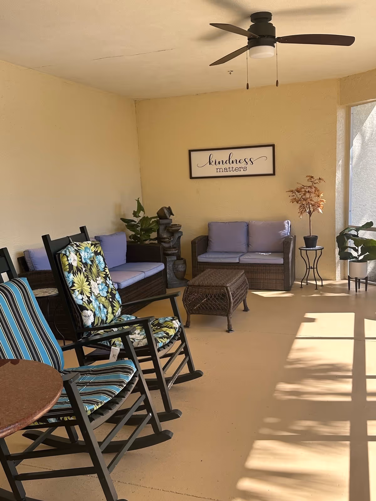 A cozy indoor seating area with two cushioned wicker loveseats, two cushioned rocking chairs with floral and striped patterns, a wicker coffee table, potted plants, a small side table, and a ceiling fan. A framed sign on the wall reads 'kindness matters'. Sunlight casts shadows on the floor from a window or screened area.
