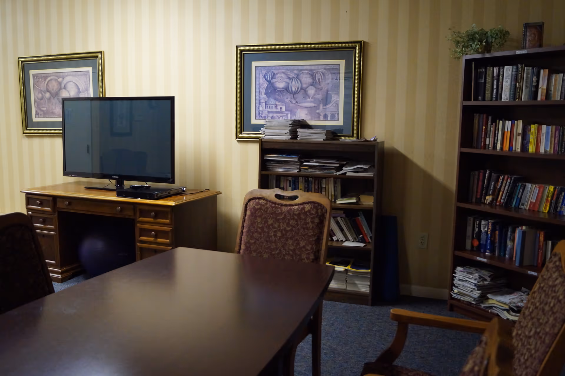 A cozy room with a dark wooden table and upholstered chairs in the foreground. Against the wall, there is a wooden desk with a flat-screen TV on top, and a purple exercise ball underneath. Two framed pictures hang on the striped wallpapered wall. Next to the desk, there is a bookshelf filled with books and magazines, and another larger bookshelf filled with books and some plants on top.