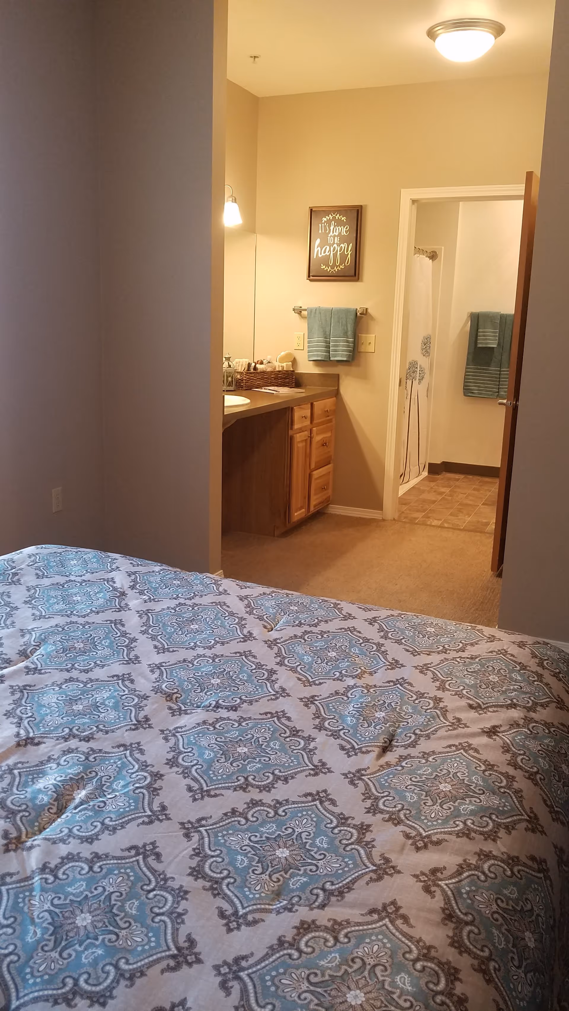 View from a bedroom into a bathroom area. The foreground shows a bed with a patterned blue and beige comforter. The bathroom area has a wooden vanity with a sink, a towel rack with blue towels, a framed sign on the wall that reads 'It's time to be happy,' and a shower with a curtain featuring a floral design.