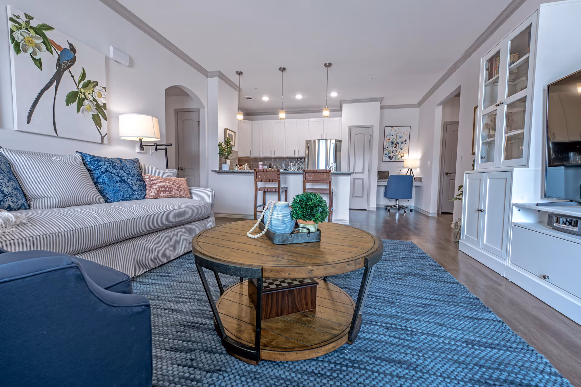 A modern living room with a striped sofa adorned with blue and pink cushions, a round wooden coffee table with decorative items, a blue armchair, and a blue patterned rug. The room opens into a kitchen area with white cabinets, stainless steel refrigerator, and three pendant lights hanging over a breakfast bar with two chairs. A small desk with a blue chair and a lamp is visible in the background.