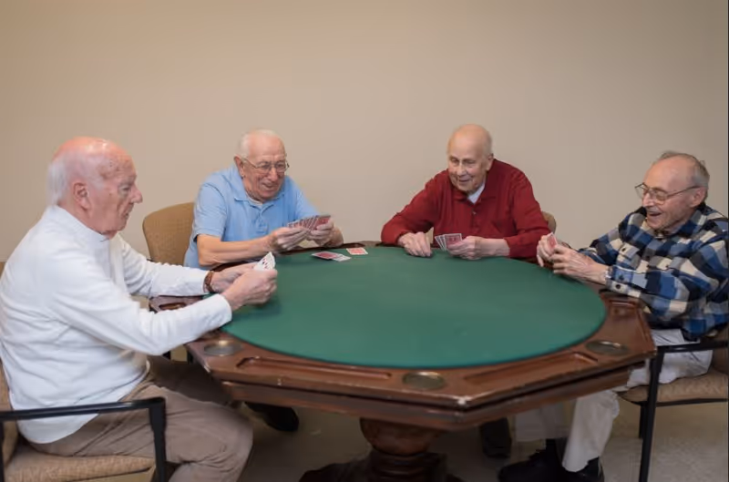 Four elderly men sitting around a green felt card table playing cards in a room with beige walls.