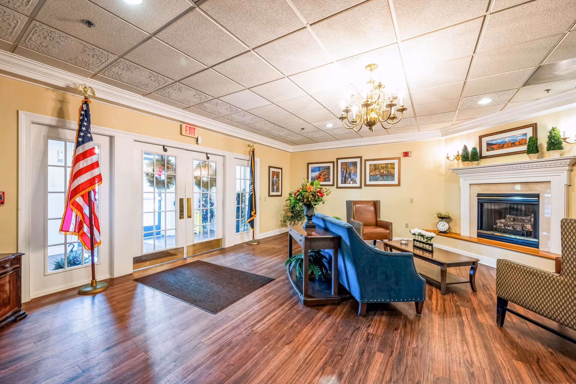 A bright and welcoming senior living facility lobby with wooden flooring, a blue upholstered sofa, brown leather and patterned chairs, a wooden coffee table, and a fireplace with decorative plants on the mantel. The walls are adorned with framed artwork, and there are two flags near the glass entrance doors with wreaths visible outside. A chandelier hangs from the ceiling providing warm lighting.