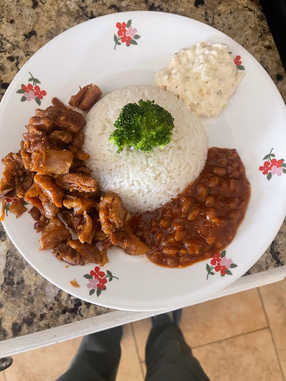 A plate of food with white rice topped with a piece of broccoli, cooked beans in a reddish sauce, a serving of cooked meat, and a portion of creamy salad, all on a white plate with red and pink floral designs, placed on a granite countertop.