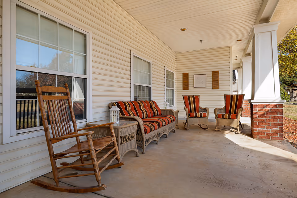 A covered outdoor porch area with a wooden rocking chair, a wicker loveseat, two wicker armchairs with red and orange striped cushions, and a small wicker side table with a white lantern on it. The porch has white siding, large windows, and white columns with brick bases. Trees and grass are visible in the background.