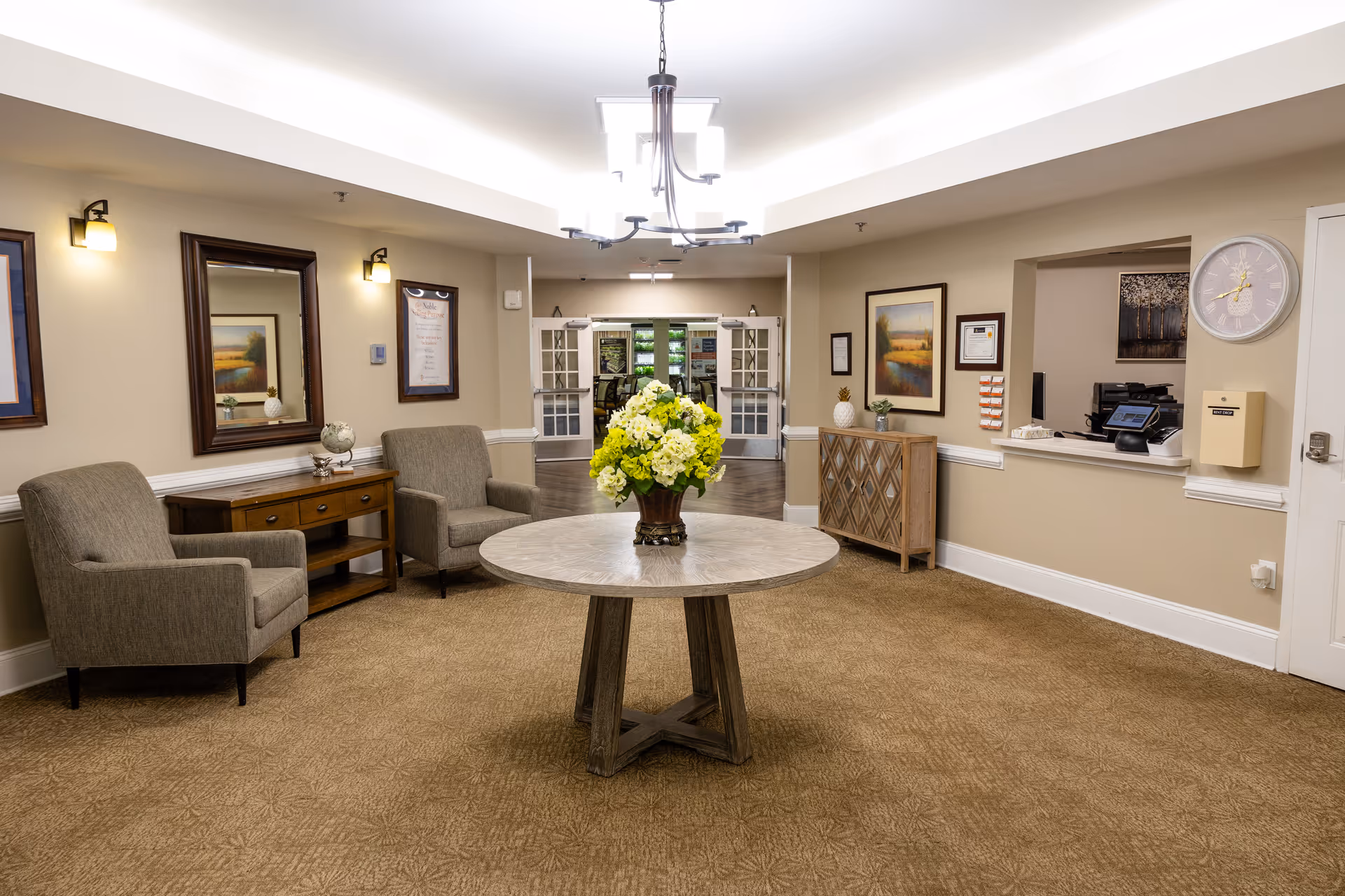 A well-lit senior living facility lobby area with a round wooden table in the center holding a vase of yellow and white flowers. Two upholstered armchairs flank a wooden console table with a globe and decorative items on the left side. On the right side, there is a wooden cabinet with framed artwork above it and a reception window with a clock on the wall. Double glass doors lead to another room in the background.