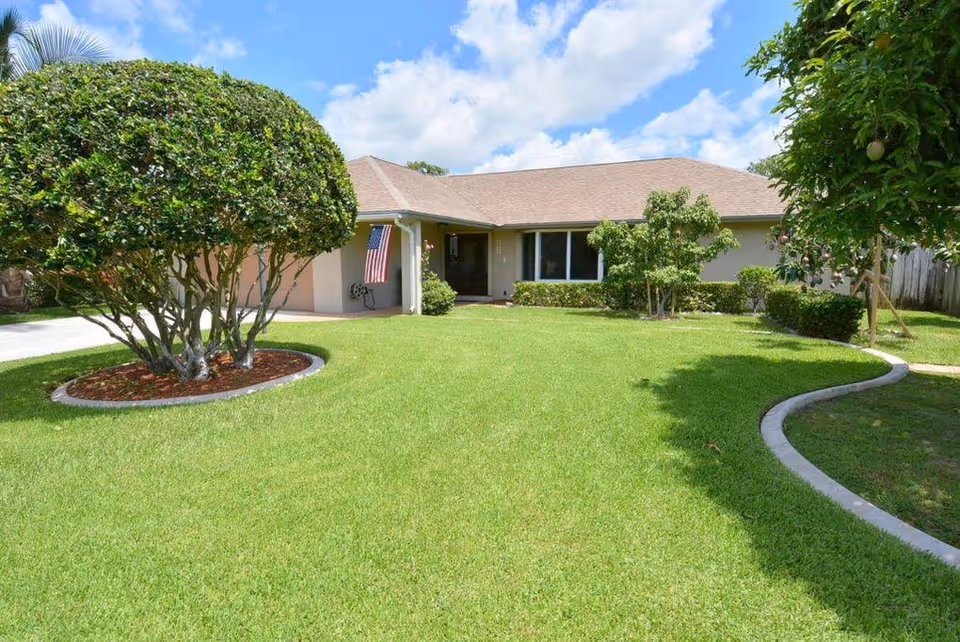 Single-story residential building with a well-maintained front lawn, trimmed bushes, and a clear blue sky with some clouds. An American flag is displayed near the entrance.