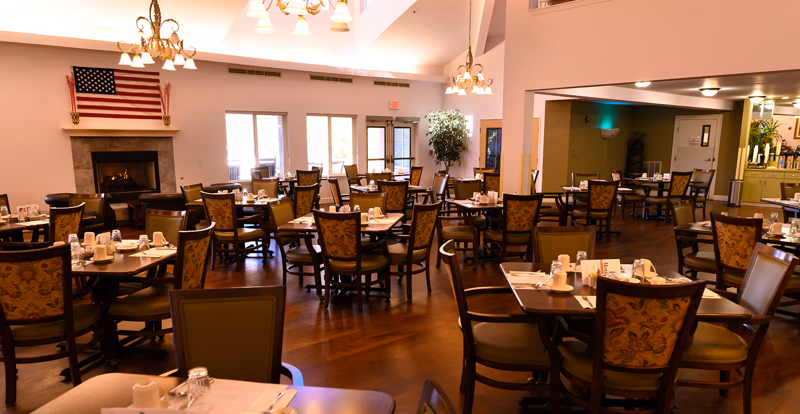 A spacious dining room with multiple wooden tables and chairs arranged neatly. Each table is set with white cups, glasses, and napkins. There is a fireplace with an American flag above it on the left side of the room. Large windows and glass doors allow natural light to enter. The room has warm lighting from chandeliers hanging from the ceiling.