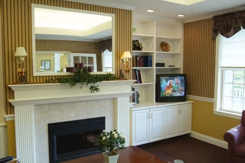 A cozy living room area with a fireplace, a large mirror above it, and built-in white shelves and cabinets. The shelves hold books and decorative items. A small television is on the cabinet showing a colorful image. There is a window with a valance letting in natural light and part of a maroon armchair is visible.