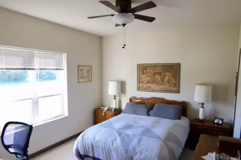 A sunlit bedroom with a bed, two nightstands and lamps, a ceiling fan, and a window.