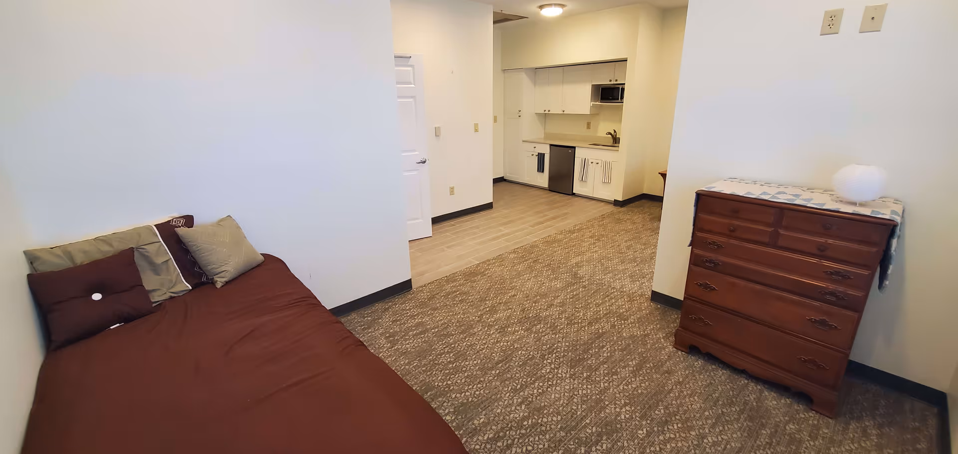 Studio-style bedroom with a single bed on the left, a wooden dresser on the right, and a small kitchenette visible at the back.