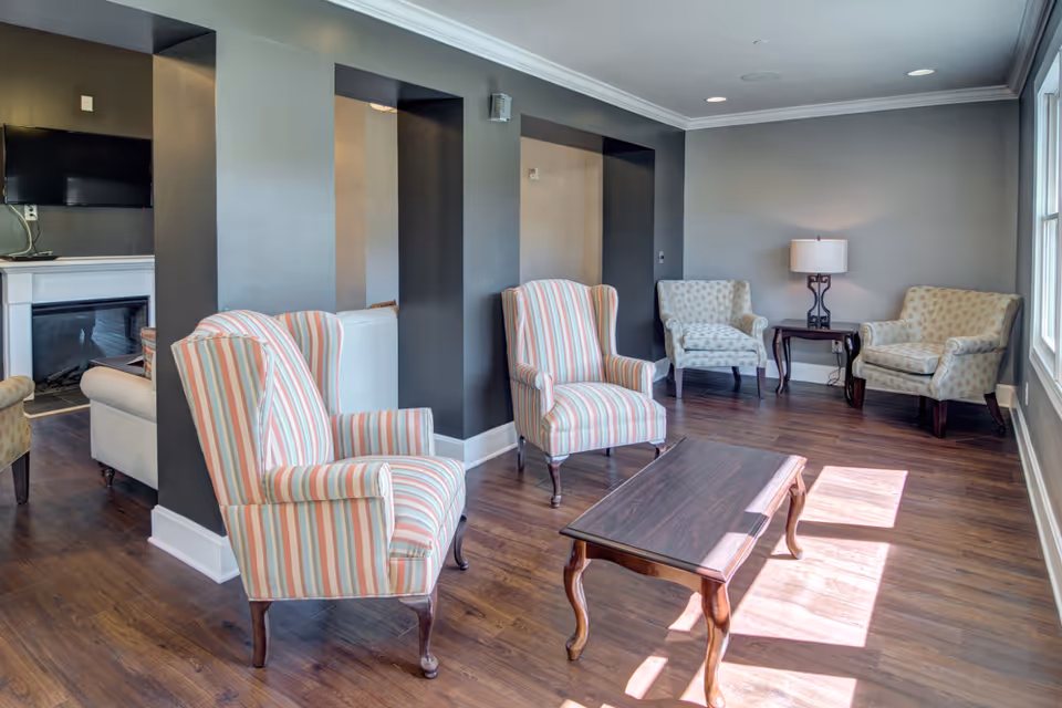 A bright common lounge with striped and patterned armchairs around a wooden coffee table on hardwood floors, with a TV and fireplace visible in an alcove.