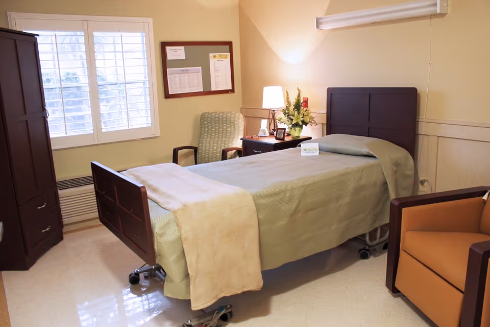 A furnished resident bedroom with a single hospital-style bed, bedside table with lamp and flowers, an armchair, and a wardrobe.