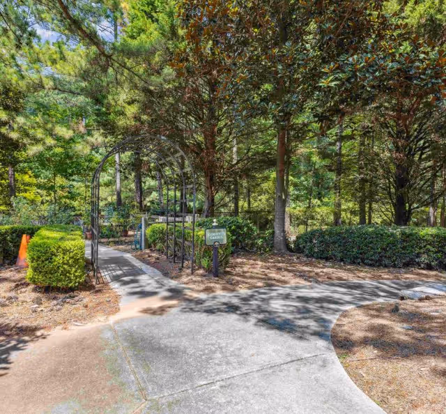 A paved pathway in a garden area surrounded by trees and bushes. There is a metal archway over the path and a small sign that reads 'Resident Gardens'. The scene is bright and sunny with shadows from the trees.