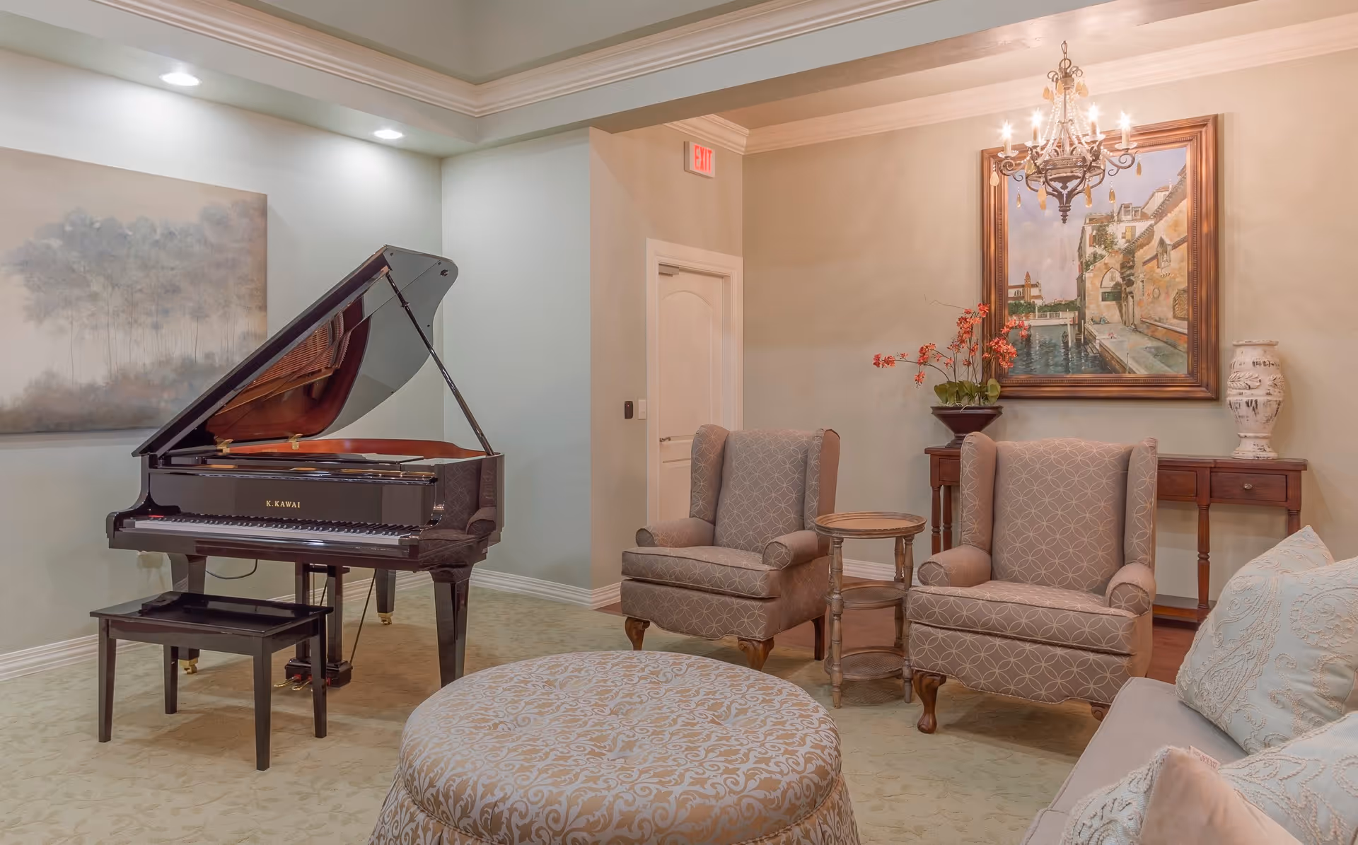 A cozy living room area with a black grand piano on the left, two patterned armchairs with a small round wooden table between them, a large round upholstered ottoman in the center, and a sofa with decorative pillows on the right. The walls are decorated with a large painting above a wooden console table with a vase of flowers and a ceramic vase. A chandelier hangs from the ceiling, and there is soft, warm lighting throughout the room.