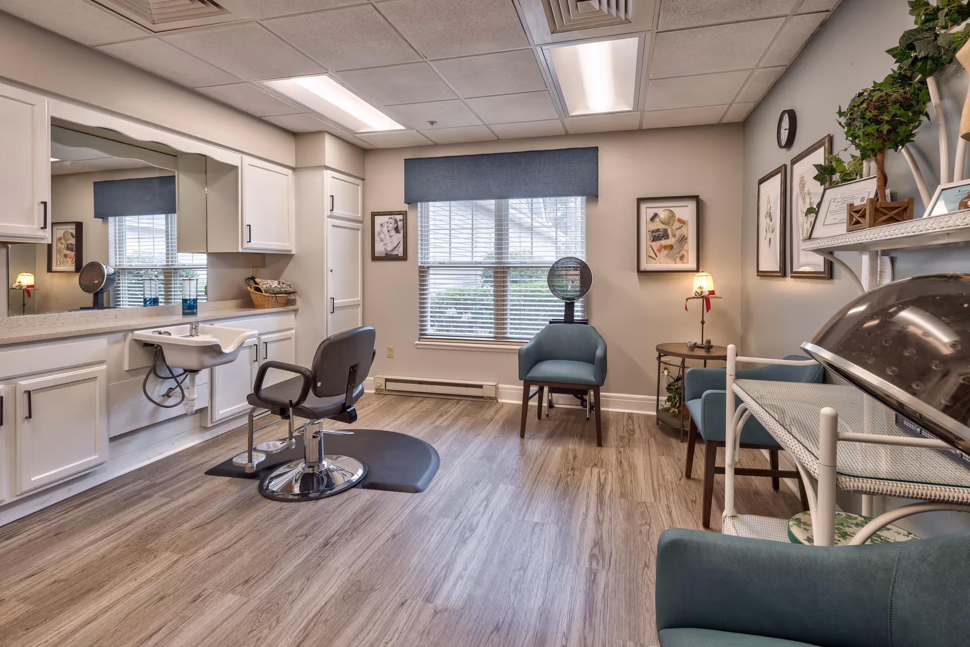 A bright and clean salon room with a hair washing station, a salon chair, and a hair dryer chair. The room has wood flooring, white cabinetry with a large mirror, a window with blinds and a blue valance, and several framed pictures and plants on the walls and shelves.