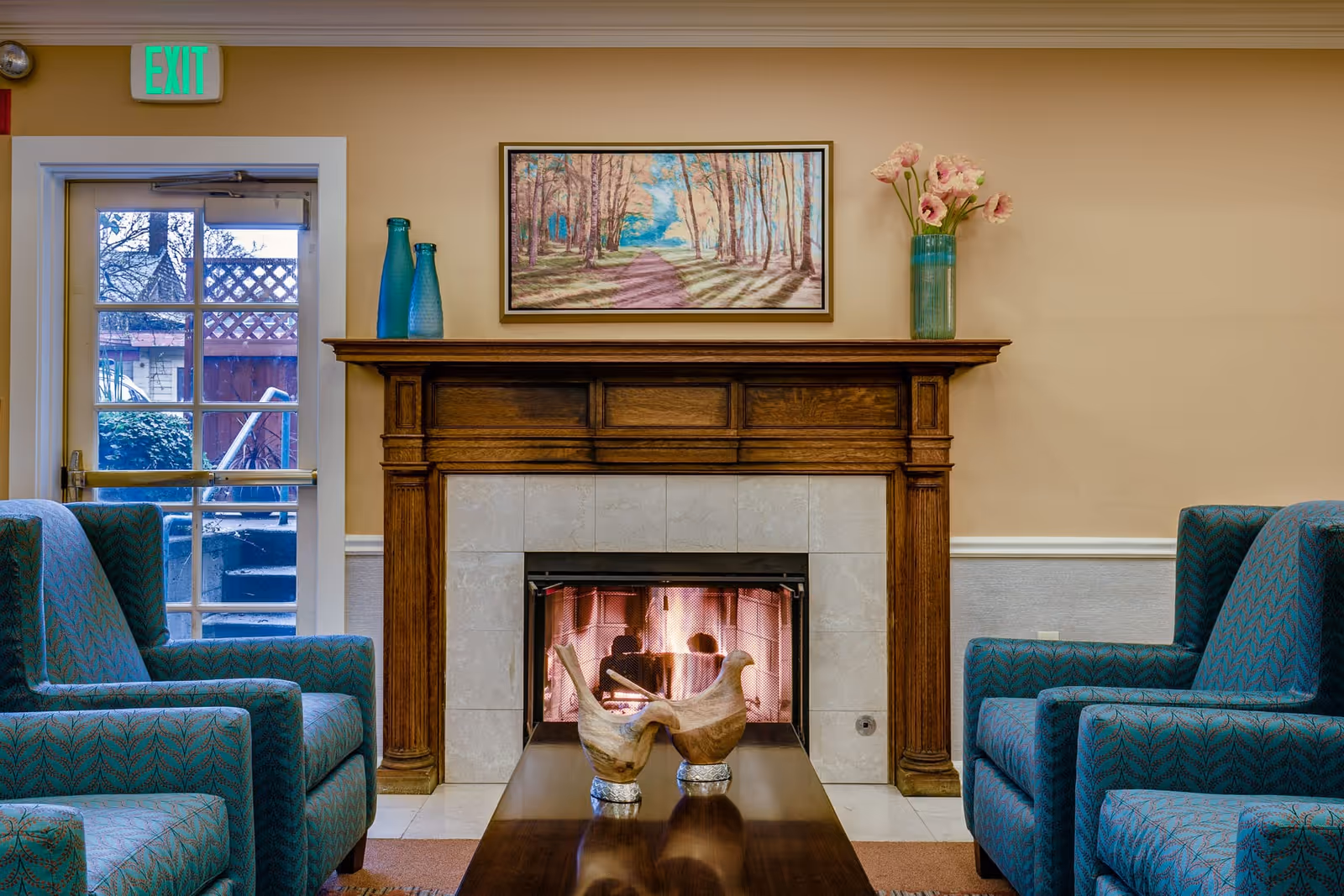 Cozy common room with a lit fireplace and wooden mantel, artwork above, and teal upholstered chairs facing a coffee table.