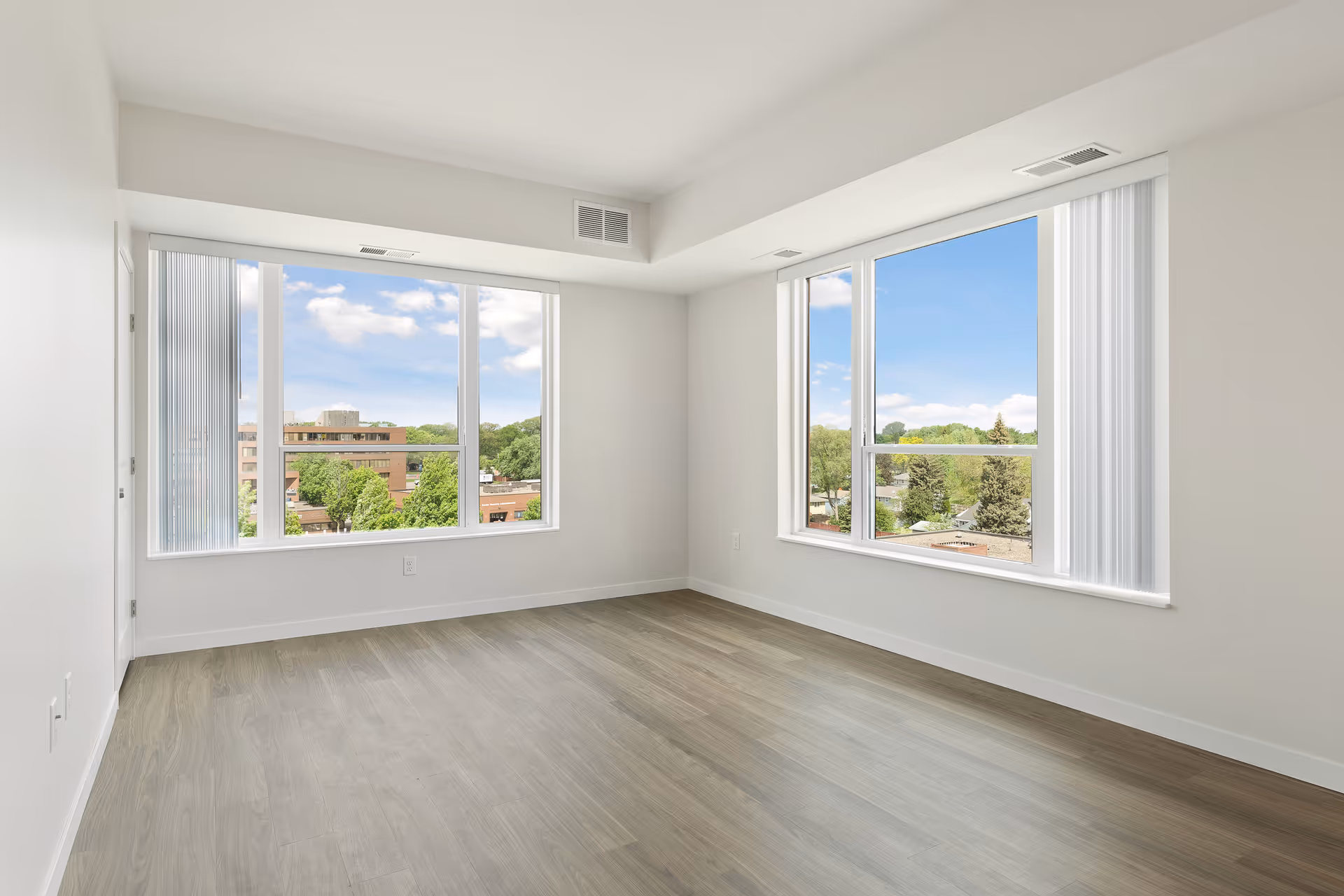 Empty room with light wood flooring and two large windows showing a view of trees and buildings outside under a blue sky with some clouds. The walls and ceiling are white, and there are vertical blinds partially covering the windows.