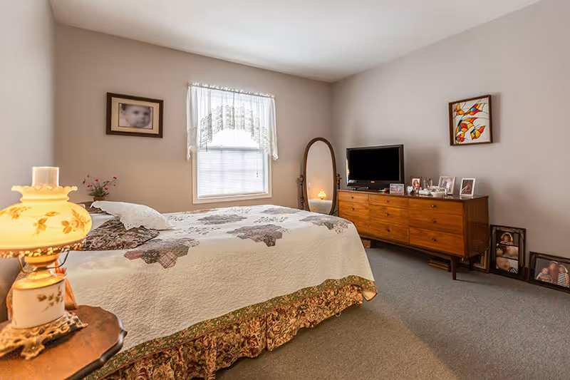Sunlit bedroom with a quilted bed, wooden dresser topped with a TV and framed photos, a floor mirror, and a bedside lamp.