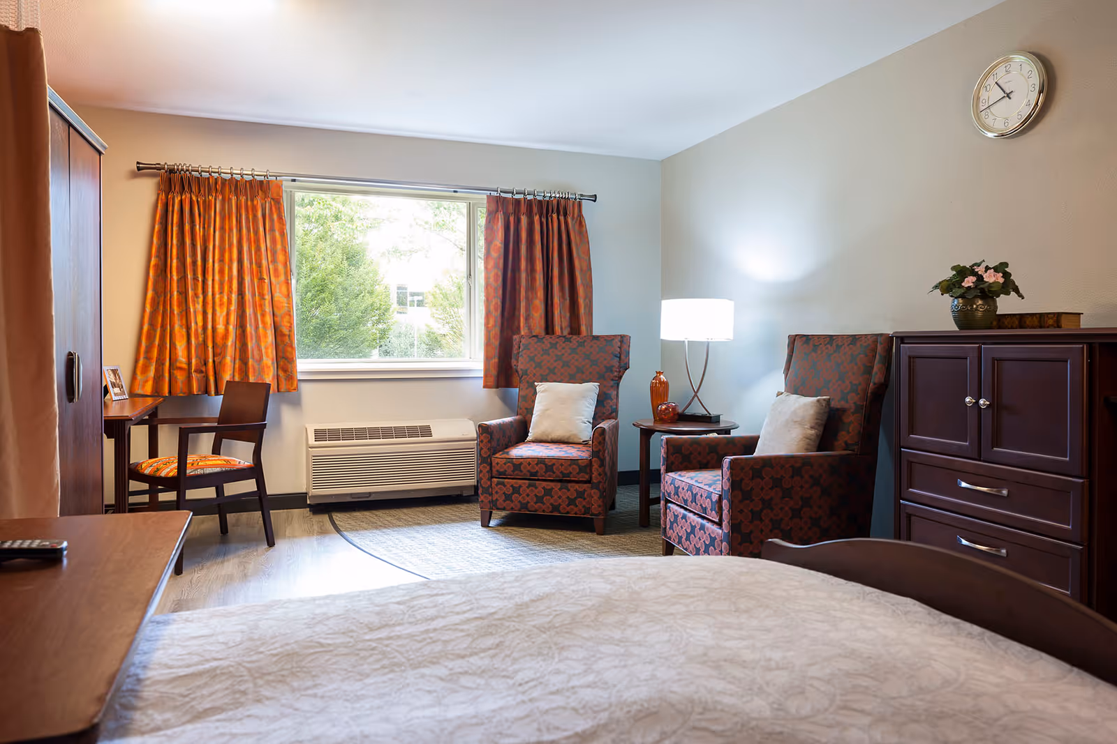 A cozy bedroom in a senior living facility featuring a bed with a white patterned bedspread in the foreground, two patterned armchairs with cushions, a wooden dresser with a plant and books on top, a small side table with a lamp, a window with orange patterned curtains, and a wooden chair and desk near the window.