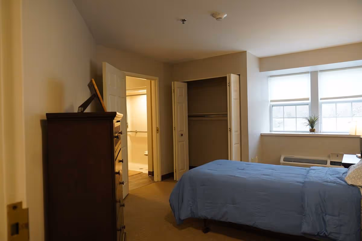 A bedroom in a senior living facility with a bed covered in a blue comforter, a wooden dresser, an open closet with bi-fold doors, and a window with white blinds letting in natural light. The door to a bathroom is open, showing grab bars inside.