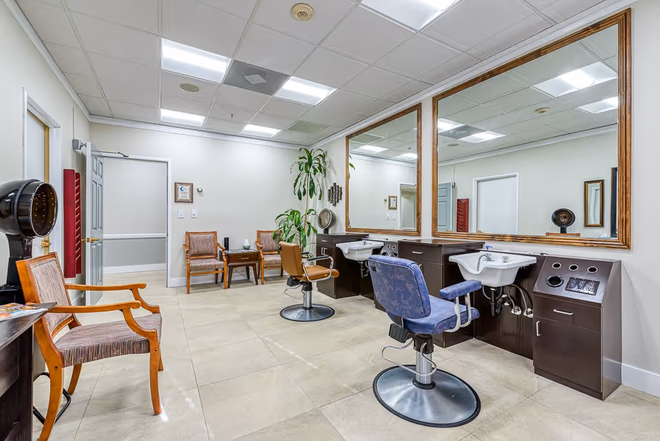 Interior view of a salon area in a senior living facility with two salon chairs in front of large mirrors and sinks, a vintage hair dryer, wooden chairs, a small table, and potted plants.