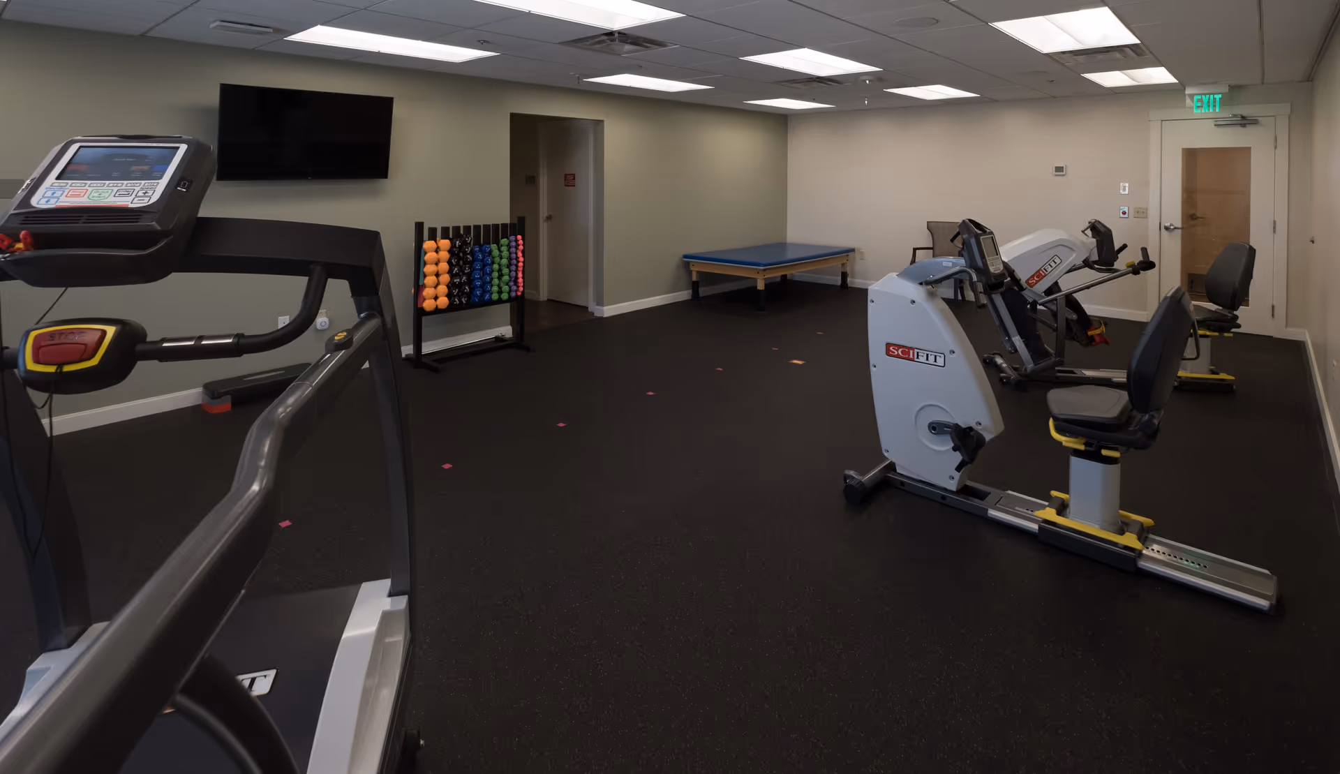 A fitness room with exercise equipment including a treadmill, two recumbent exercise bikes, a rack of colorful dumbbells, a blue padded exercise table, and a wall-mounted television. The room has a black rubber floor, light-colored walls, and a door with an exit sign above it.
