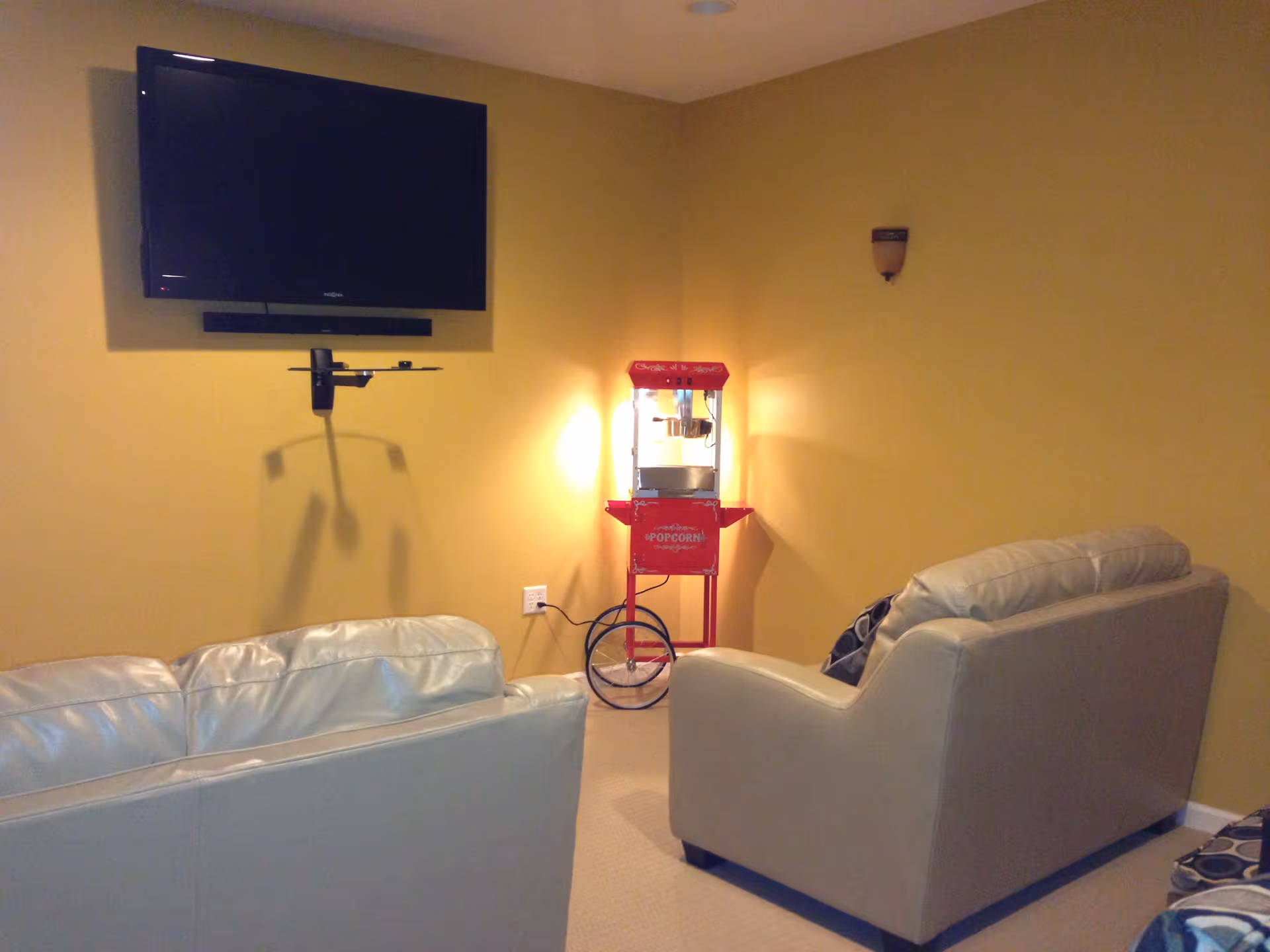 A cozy living room with two light-colored leather sofas facing a wall-mounted flat screen TV. In the corner, there is a red vintage-style popcorn machine illuminated by a wall sconce on a yellow wall.