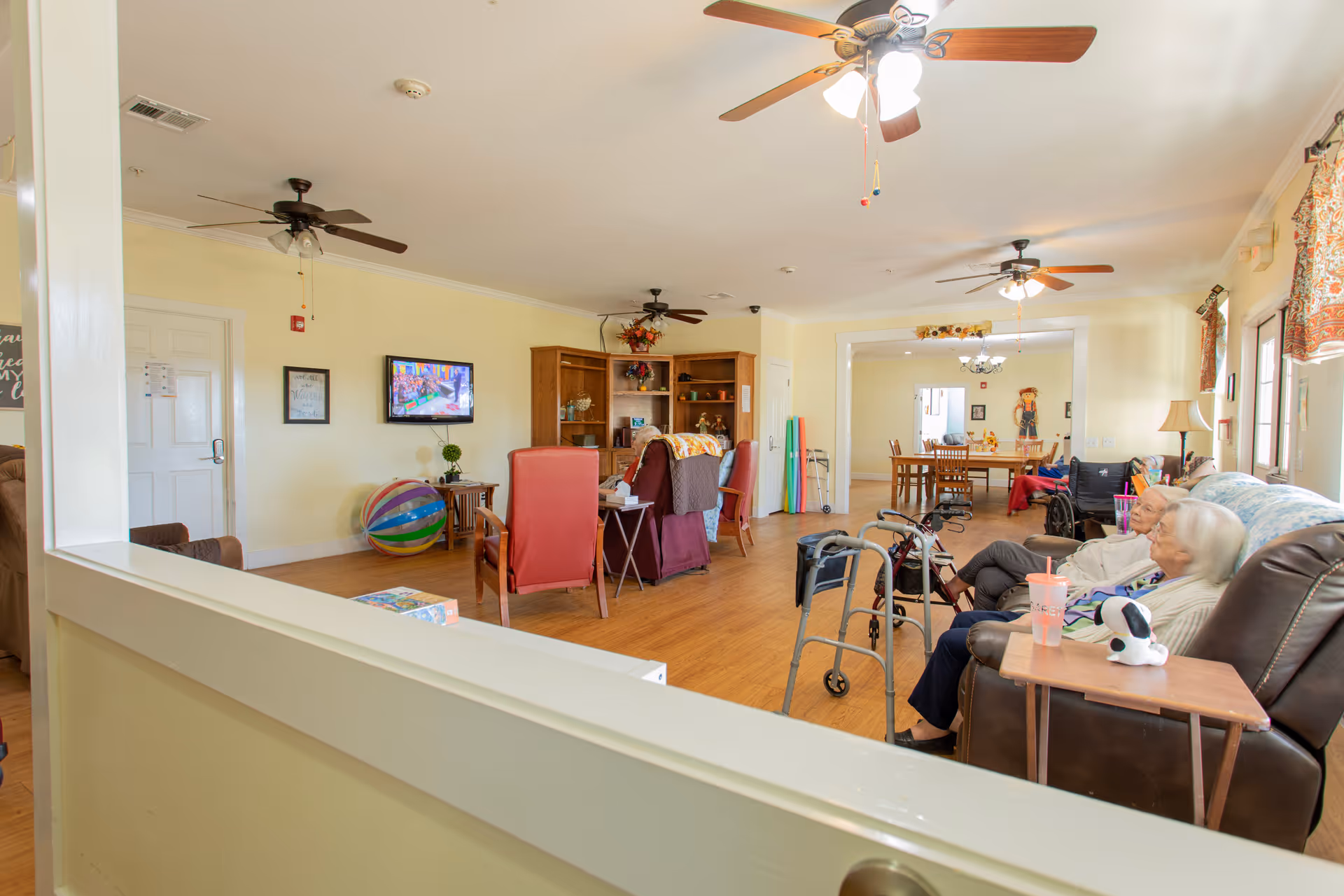 A bright and spacious common living area in a senior living facility with several ceiling fans, comfortable chairs, a television mounted on the wall, and a few elderly residents seated and relaxing. The room has wooden flooring, light yellow walls, and large windows with patterned curtains allowing natural light to enter. In the background, there is a dining area with a table and chairs.