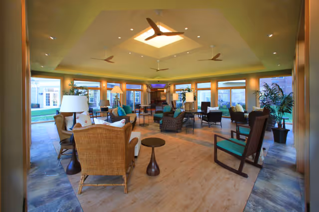 A spacious and well-lit living room area with multiple seating options including wicker and cushioned chairs, small tables, and floor lamps. The room features large windows allowing natural light to enter, ceiling fans, and a skylight in the center of the ceiling. There are plants placed around the room, and the floor is a combination of wood and stone tiles.