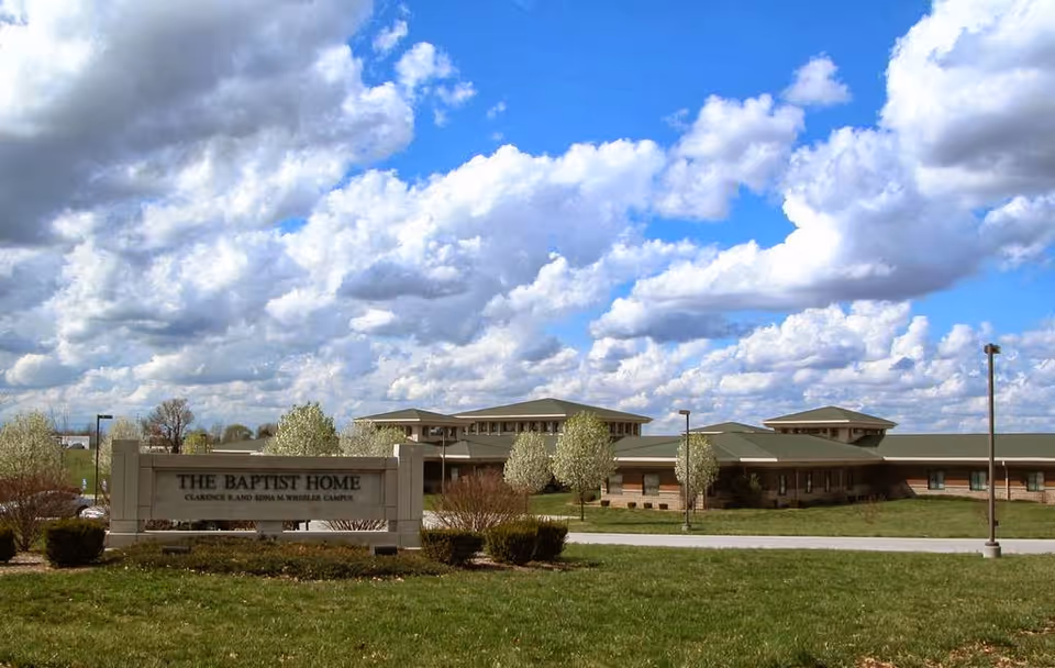 Exterior view of The Baptist Home, Clarison Bland Edna M. Wheeler Campus, showing a single-story building with a green roof under a partly cloudy blue sky. The foreground features a grassy area with bushes and a sign displaying the facility's name.