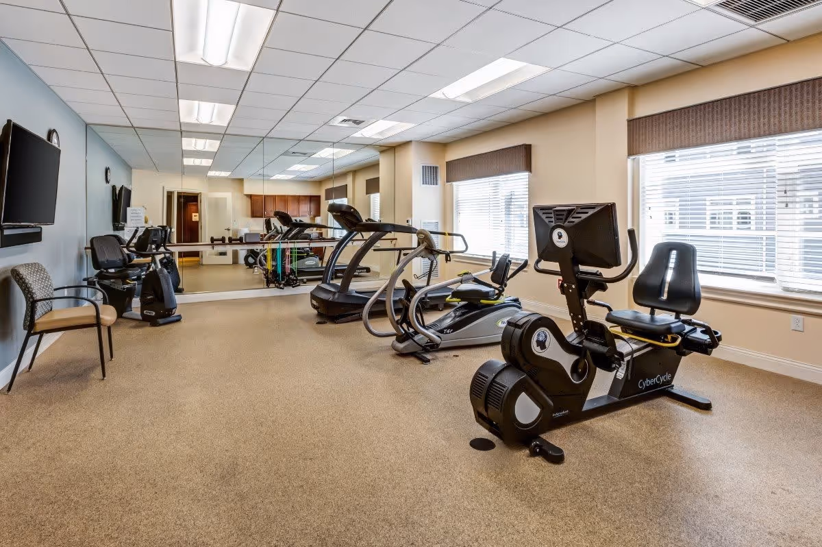 A senior living facility exercise room with various fitness equipment including a recumbent stationary bike with a screen, treadmills, and other cardio machines. The room has large windows with blinds, a wall-mounted TV, chairs, and a mirrored wall reflecting the equipment. The floor is carpeted and the ceiling has fluorescent lighting.