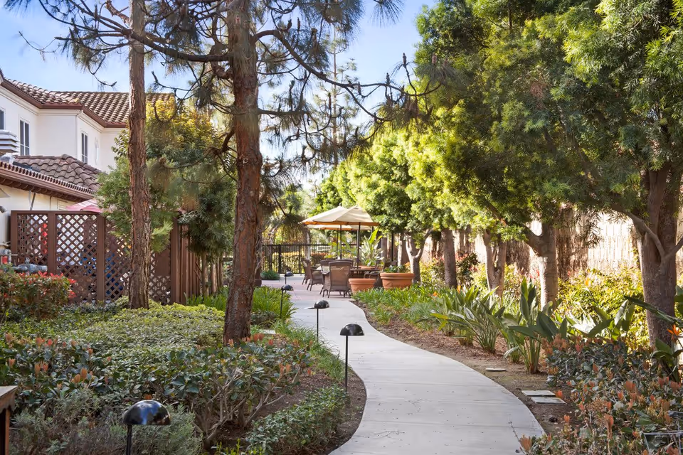 A winding concrete pathway through a landscaped garden area with trees, shrubs, and plants on both sides. There are outdoor chairs and tables with umbrellas in the background near a building with a tiled roof.