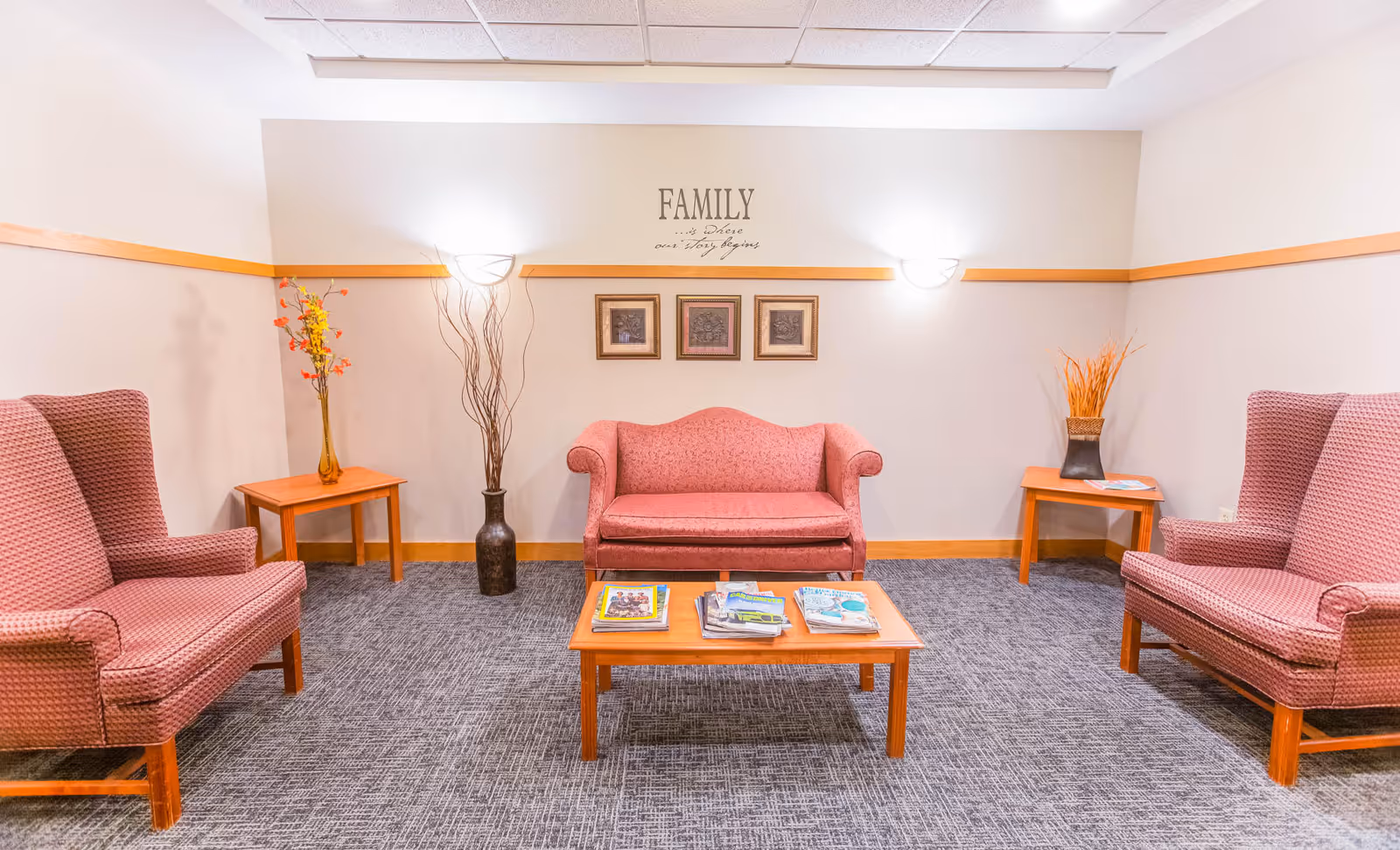 A cozy sitting area in a senior living facility with a pink upholstered loveseat and two matching armchairs arranged around a wooden coffee table with magazines. The walls are light-colored with a wooden trim, decorated with three framed pictures and a wall decal that reads 'FAMILY is where our story begins.' There are two wooden side tables with decorative vases and plants, and the floor is carpeted in a gray pattern.
