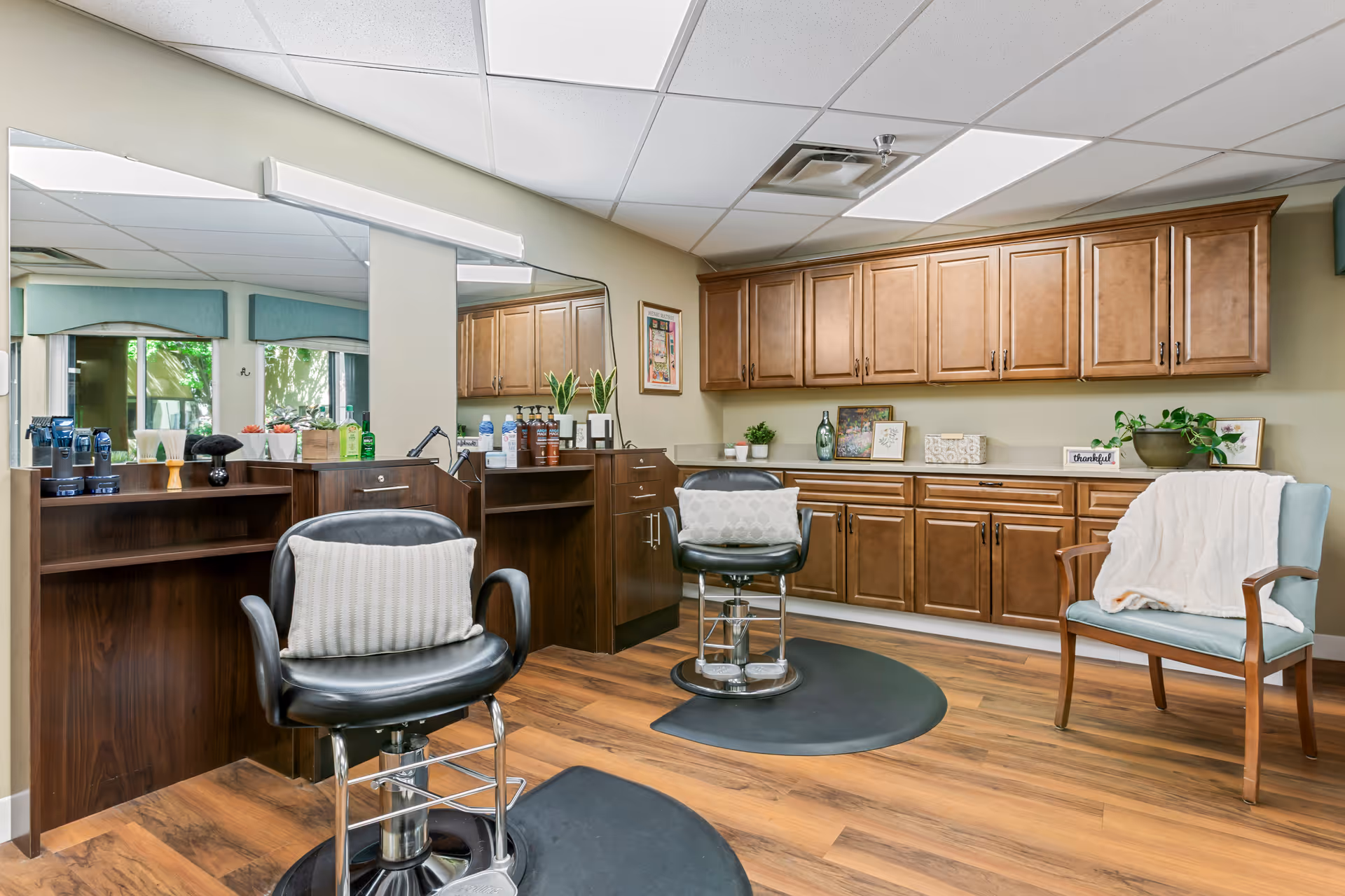 Interior view of a salon area in a senior living facility with two black salon chairs, wooden cabinetry along the wall, a large mirror, and a light blue chair with a white blanket draped over it. The floor is wood, and there are various plants and decorative items on the counters.