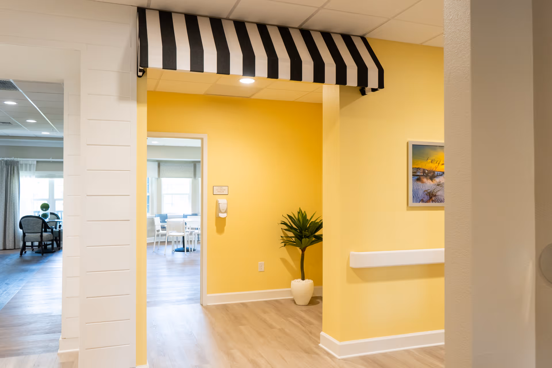Bright interior hallway with yellow walls, a black-and-white striped awning, a potted plant, and a view into a dining area.