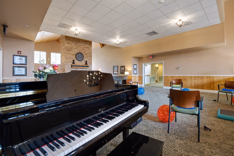 Interior view of a spacious common area in a senior living facility featuring a black grand piano in the foreground, several chairs arranged around the room, colorful exercise balls on the carpeted floor, and a stone fireplace with a clock above it in the background. The room has beige walls with wood paneling and a white tiled ceiling with light fixtures.