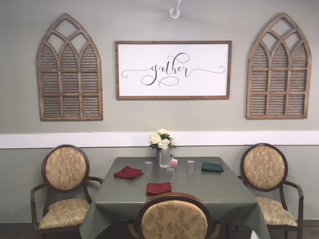 A small dining area with a square table covered with a green tablecloth, set with four chairs upholstered in a patterned beige fabric. On the table are four drinking glasses, three folded cloth napkins in red and green, and a vase with white flowers. The wall behind the table has a decorative sign that reads 'gather' in cursive script, flanked by two wooden window shutter-style wall decorations.
