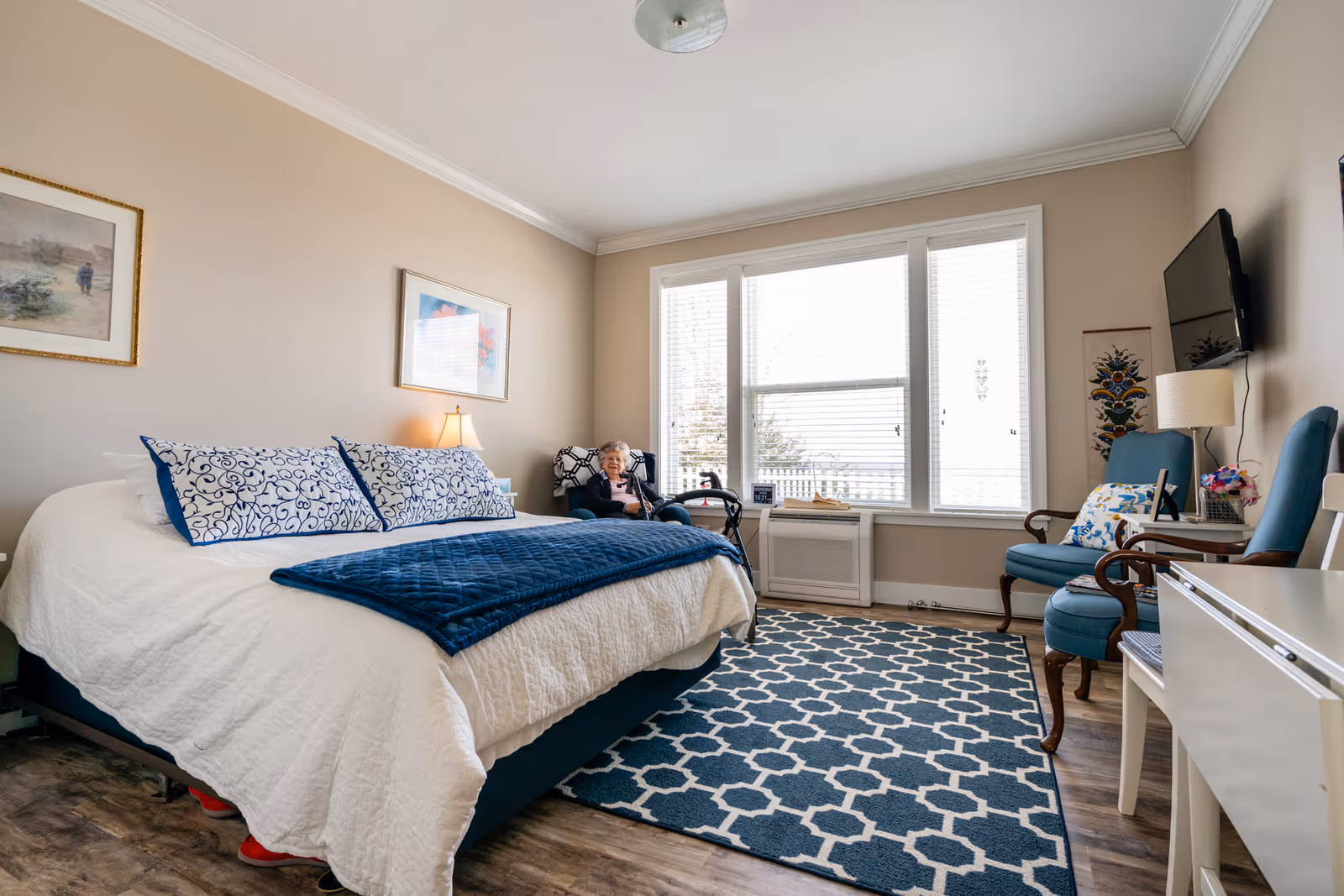 A bright and cozy bedroom in a senior living facility with a large bed covered in white and blue bedding, a patterned blue rug on the floor, two blue armchairs, a wall-mounted TV, and a large window letting in natural light. An elderly woman is sitting in one of the armchairs near the window with a walker beside her.