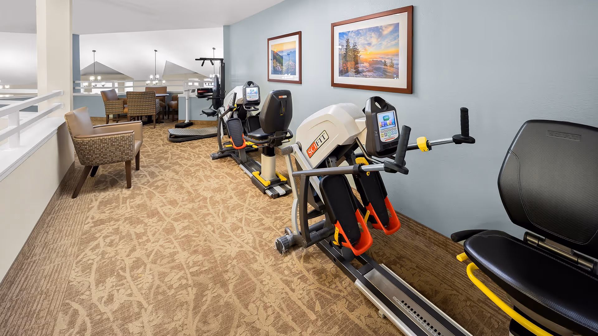 A fitness area in a senior living facility featuring exercise equipment including a SciFit machine, with a seating area and dining tables visible in the background. The walls are light blue with framed landscape pictures, and the floor is carpeted in a beige pattern.