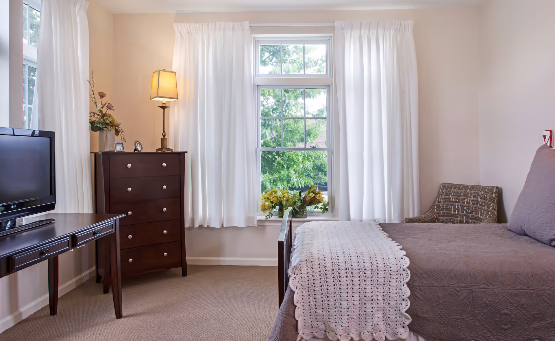 A cozy bedroom with a bed covered in a brown quilt and a white crocheted blanket at the foot. There is a patterned armchair in the corner, a wooden dresser with a lamp and decorative items on top, and a flat-screen TV on a wooden table. The room has large windows with white curtains letting in natural light and showing green trees outside.